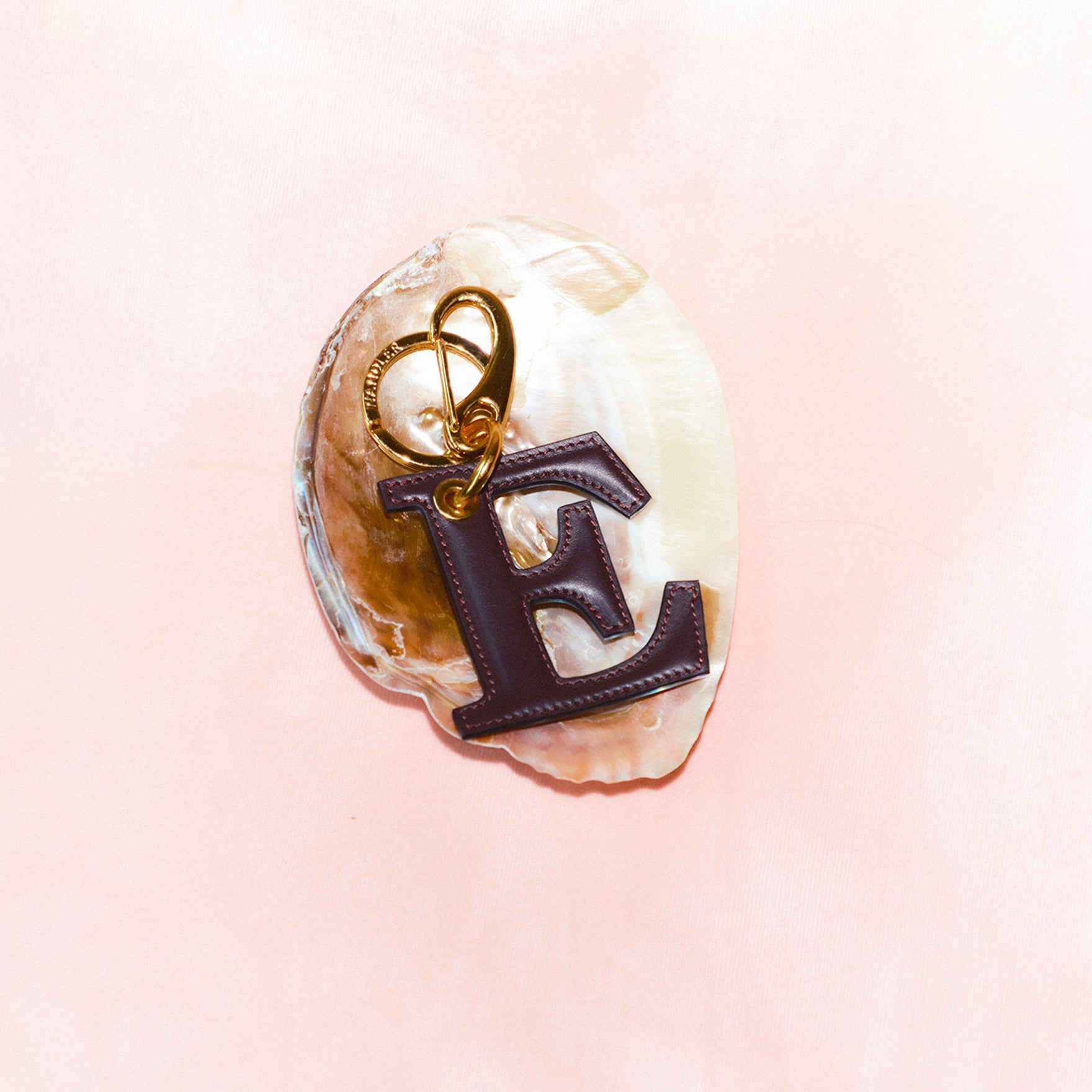 Wandler Bag Charm Letter in the color Grape, deep dark purple, made from brass and lambskin leather with golden hardware. Shown on a shell  against a soft pink background.