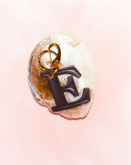 Wandler Bag Charm Letter in the color Grape, deep dark purple, made from brass and lambskin leather with golden hardware. Shown on a shell  against a soft pink background.