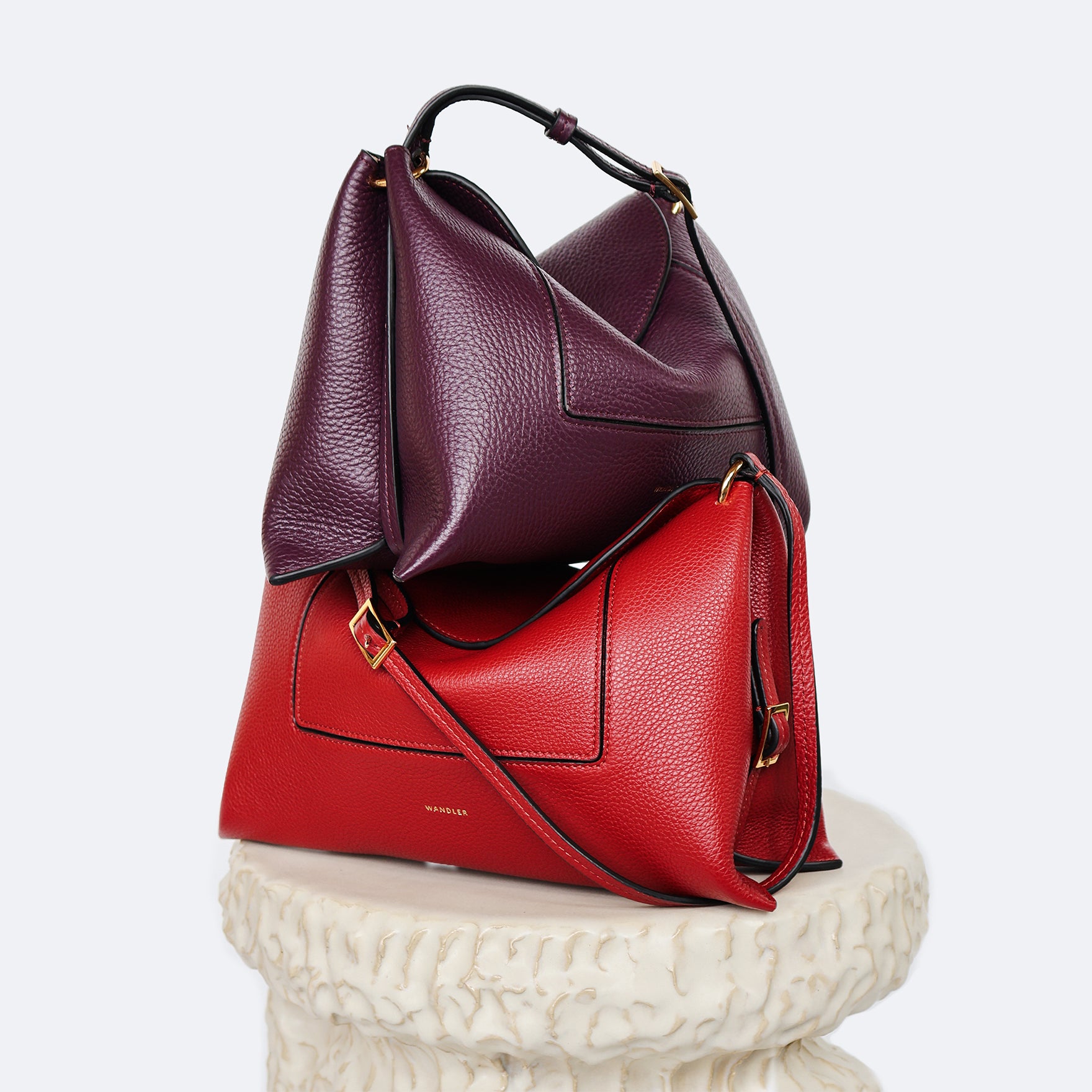 Wandler Penelope Slouch Bag in the color Scarlet Crust and Aubergine Crust, made from calf leather with golden hardware and adjustable slim strap, the bags are perpendicularly stacked on top of each other. the bags are also positioned on top of a pillar.
