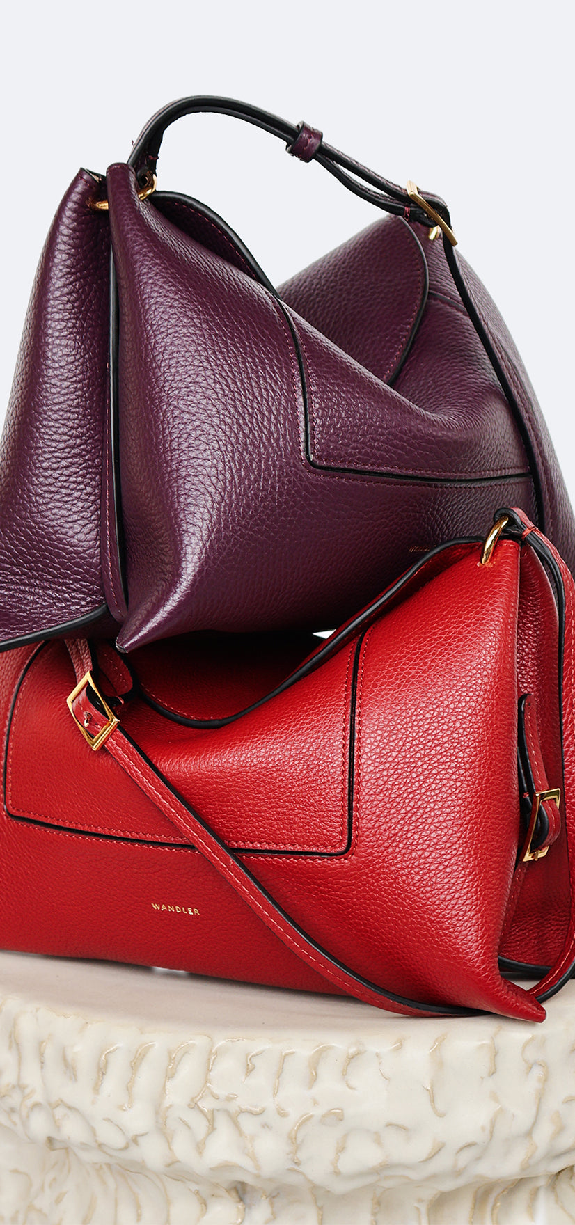 Wandler Penelope Slouch Bag in the color Scarlet Crust and Aubergine Crust, made from calf leather with golden hardware and adjustable slim strap, the bags are perpendicularly stacked on top of each other. the bags are also positioned on top of a pillar.