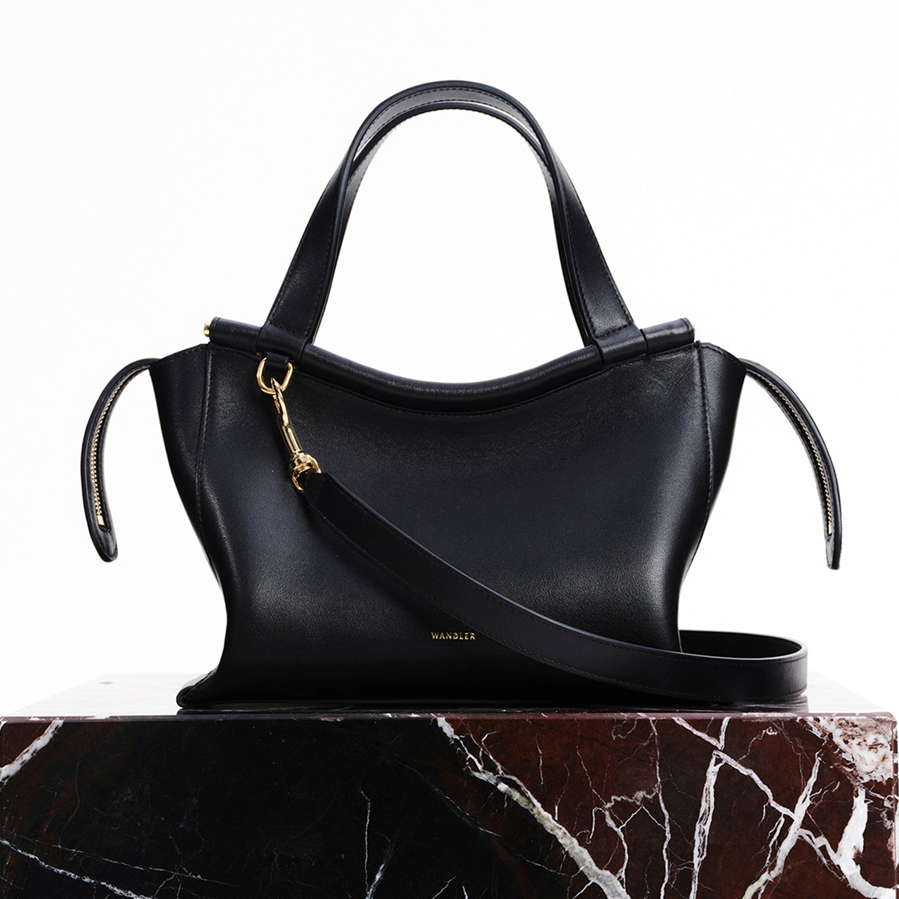 Wandler Ida Bag in the color Black, made from Italian calf leather with golden hardware and sculptural top handle, pictured on pillar.