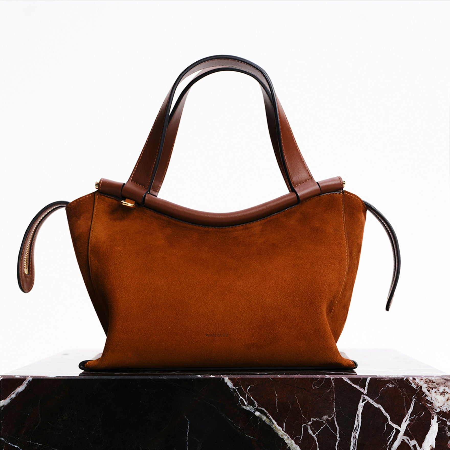 Wandler Ida Bag in the color Tan Suede, made from Italian calf leather with golden hardware and sculptural top handle, pictured on marble pillar.