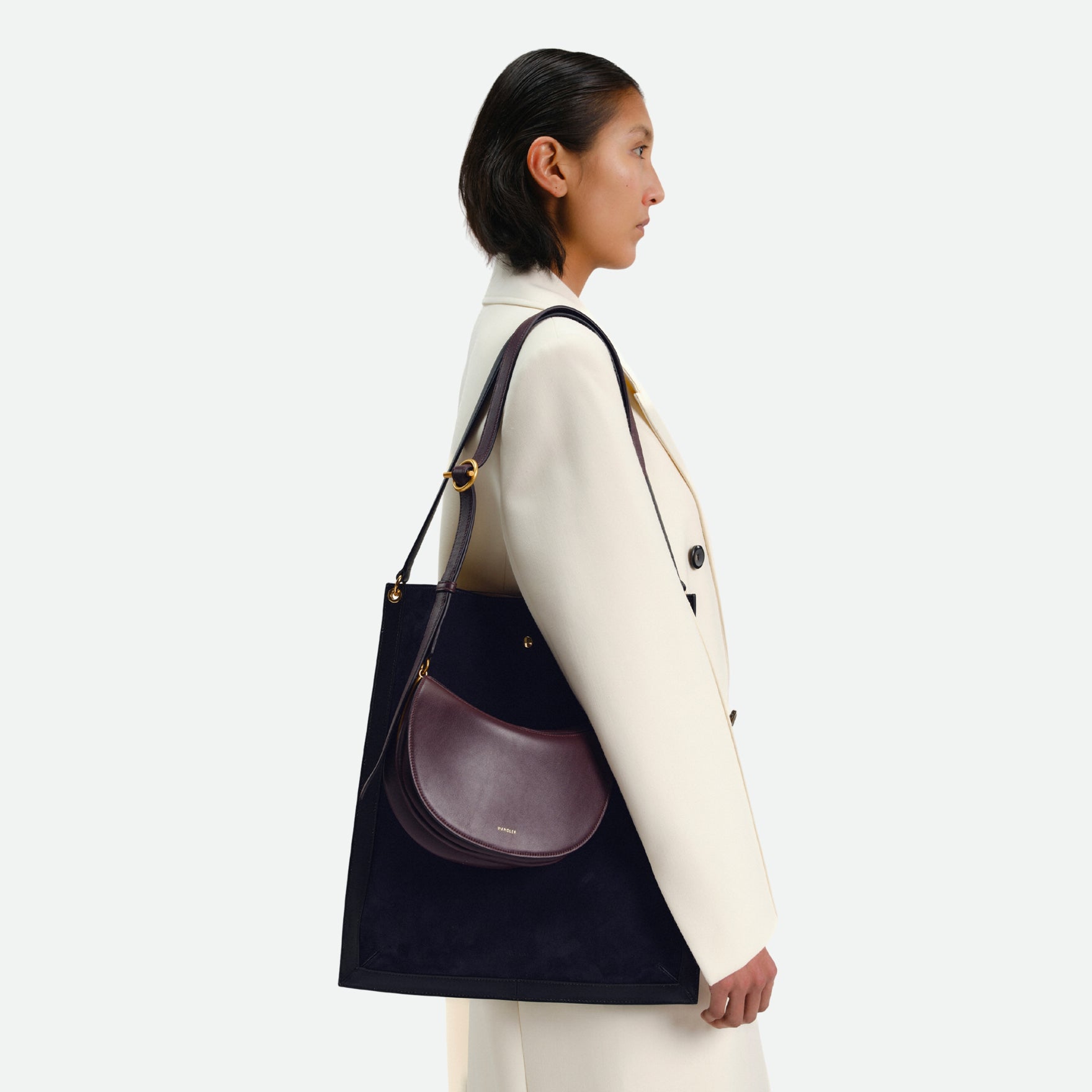 Wandler Penny Tote in the color Black Suede, made from Italian calf leather with hand-painted black edges and golden hardware, pictured on model, being worn on the shoulder together with another bag.