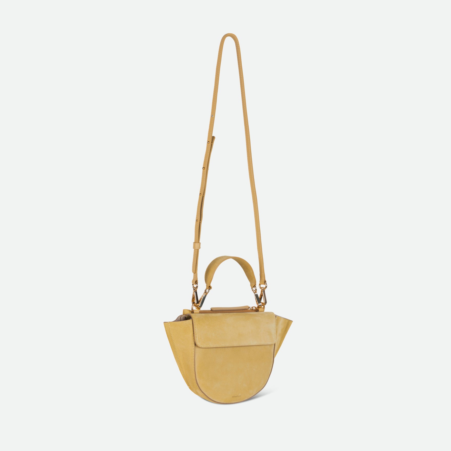 Wandler Hortensia Bag Mini in the color Sandy Suede, muted yellow suede, Italian calf leather with golden hardware, rounded silhouette and top handle. The bag is pictured with the crossbody strap extended into the air.