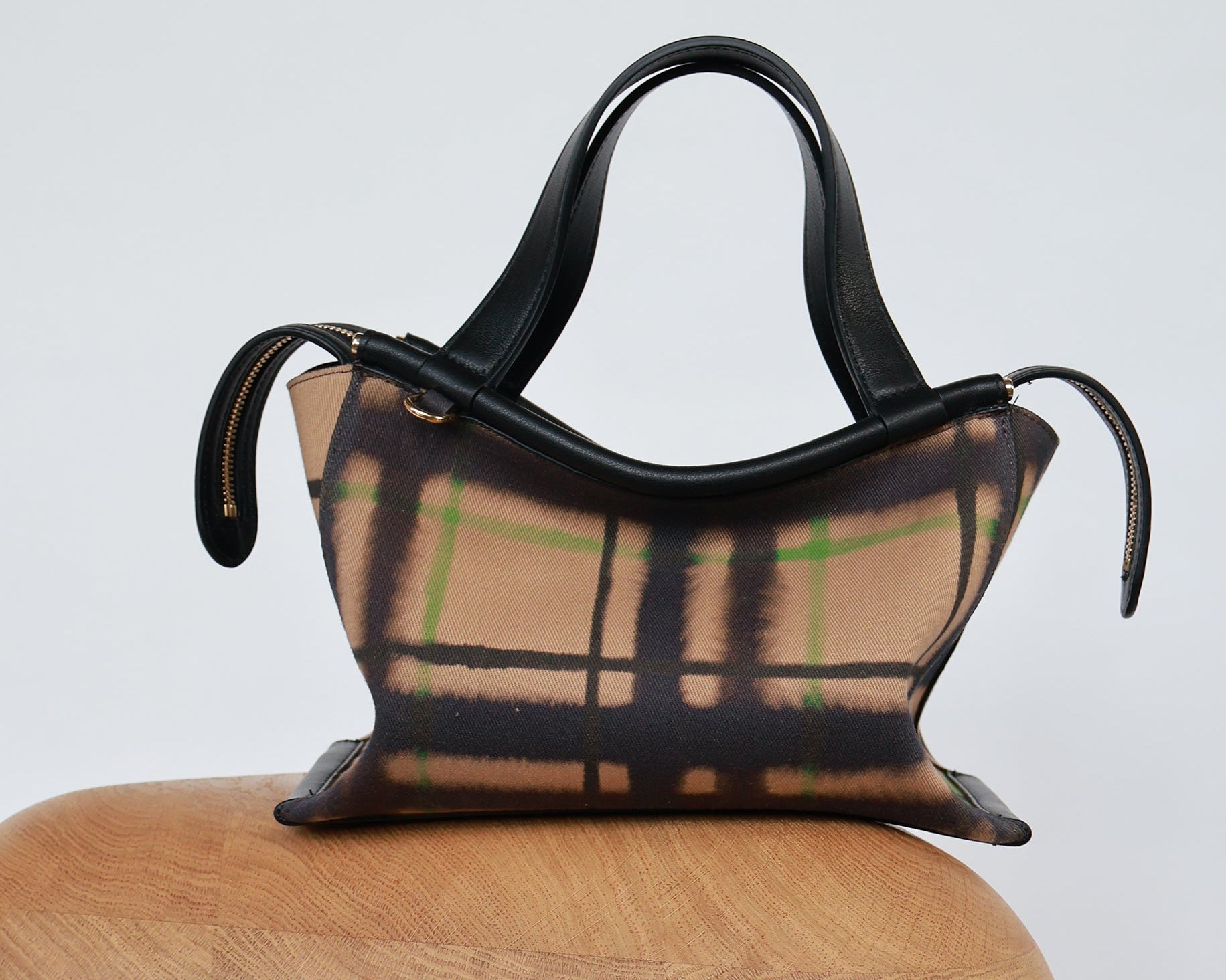 A small structured handbag with black leather handles and trim, featuring a beige plaid body with black and green lines, displayed on a wooden stool against a light gray background.