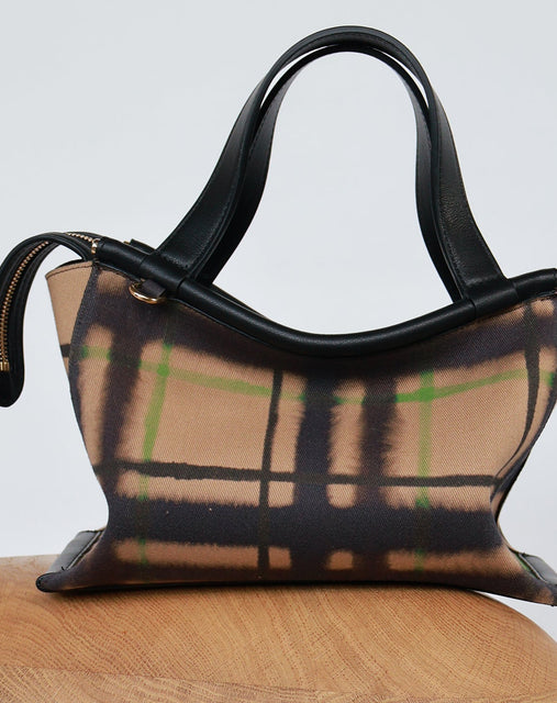 A small structured handbag with black leather handles and trim, featuring a beige plaid body with black and green lines, displayed on a wooden stool against a light gray background.