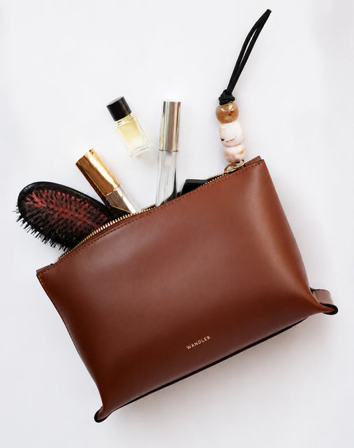 A brown leather cosmetic pouch with a gold zipper, shown flat-lay with various beauty items like a hairbrush, perfume, and lip gloss peeking out. The brand name Wandler is printed in gold foil on the front.