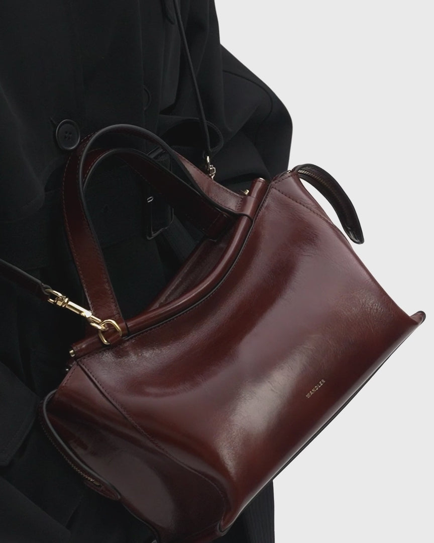 Video of Wandler Ida Bag in the color Palissander Glaze, deep dark brown with red undertones, made from Italian calf leather with golden hardware and sculptural top handle, shot on model wearing the bag in a crossbody style, displaying multiple angels as well as closeups of the bag.